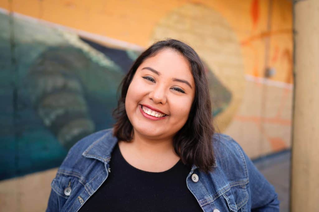 Smiling woman with dark hair in a denim jacket in front of a colorful mural.
