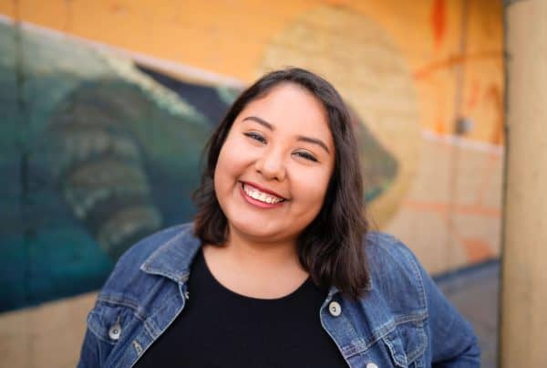 Smiling woman with dark hair in a denim jacket in front of a colorful mural.