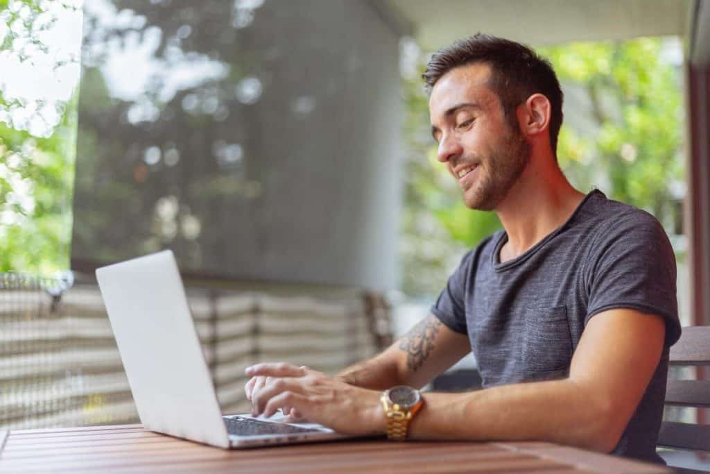 Young man working on a laptop outdoors with greenery in the background.
