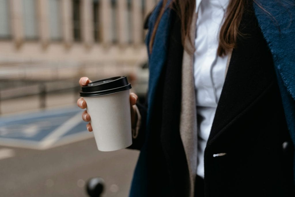 Person holding a takeaway coffee cup outdoors in an urban setting.