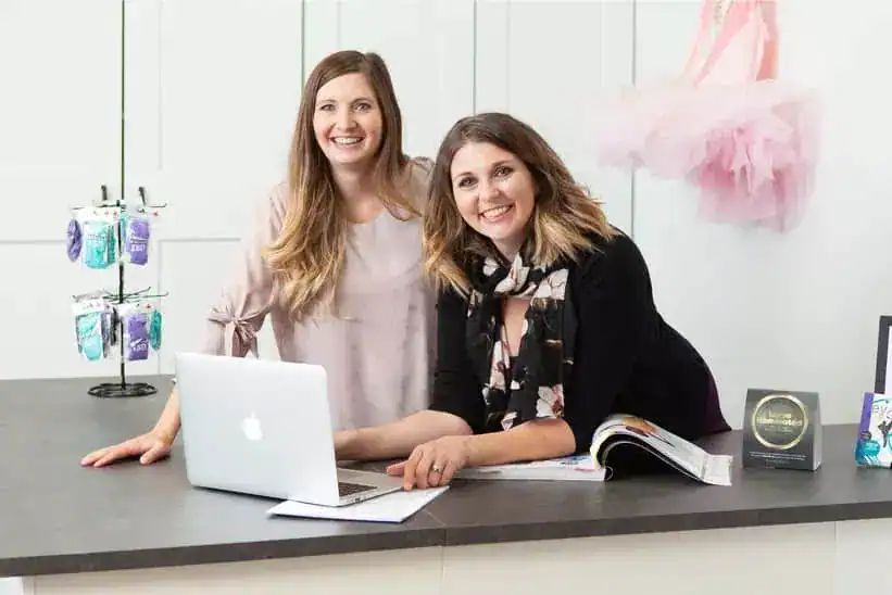 Two women smiling at a reception desk with a laptop and marketing materials.
